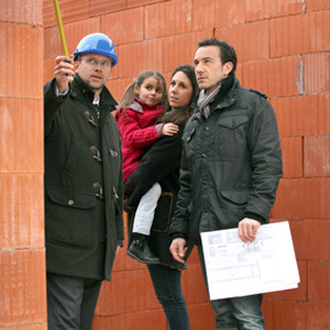 man in blue hard hat pointing with measuring tape with man, woman and child by brick wall