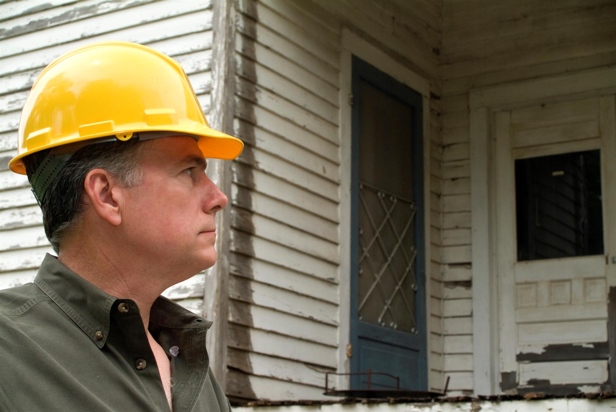 man in yellow hard hat and green shirt in front of dilapidated home